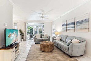 A spacious living room with a grey sofa, a television, a ceiling fan, and a large window.