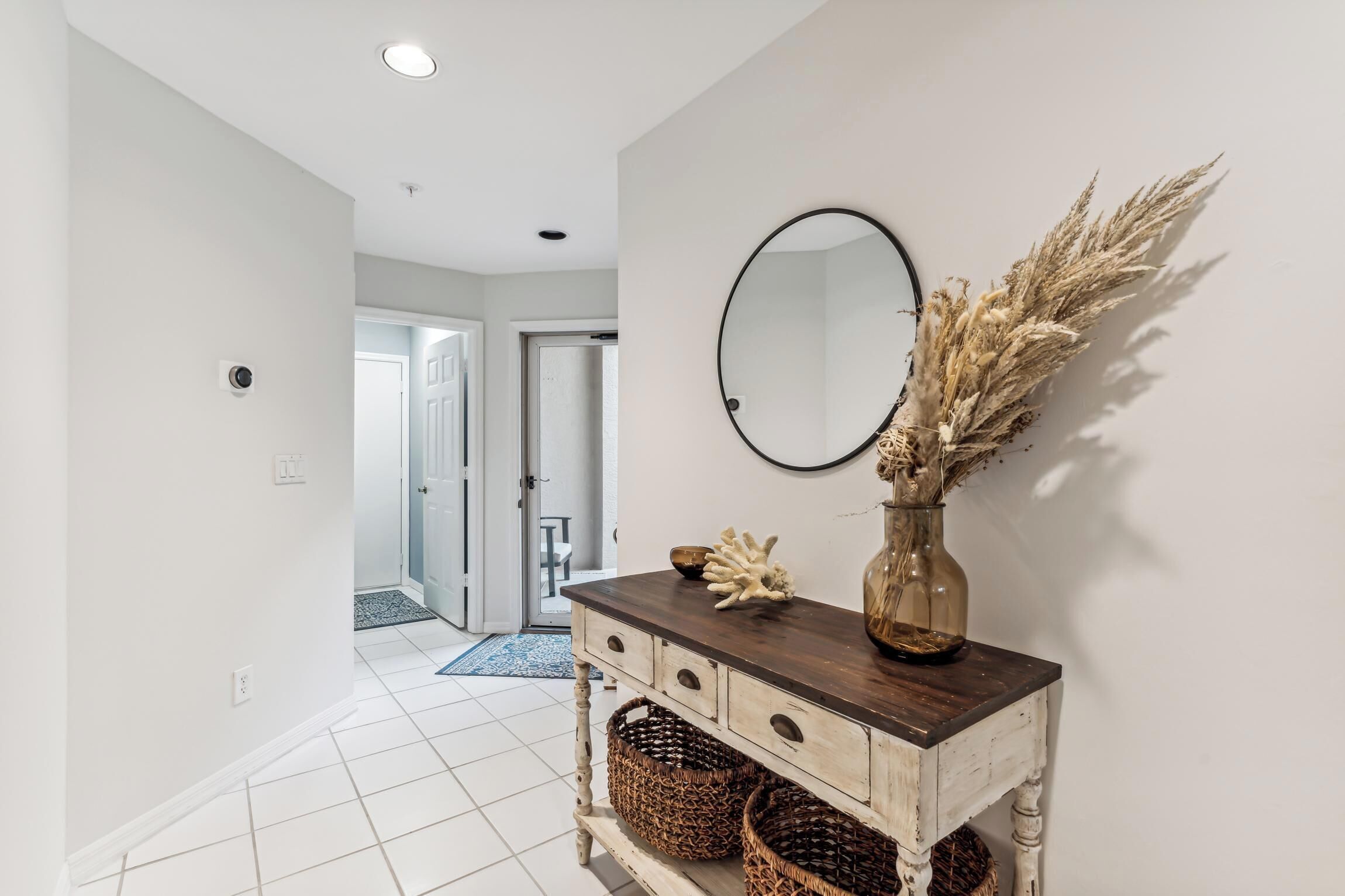 A hallway with a white tiled floor, a wooden console table with baskets, a round mirror, and a vase with dried plants.