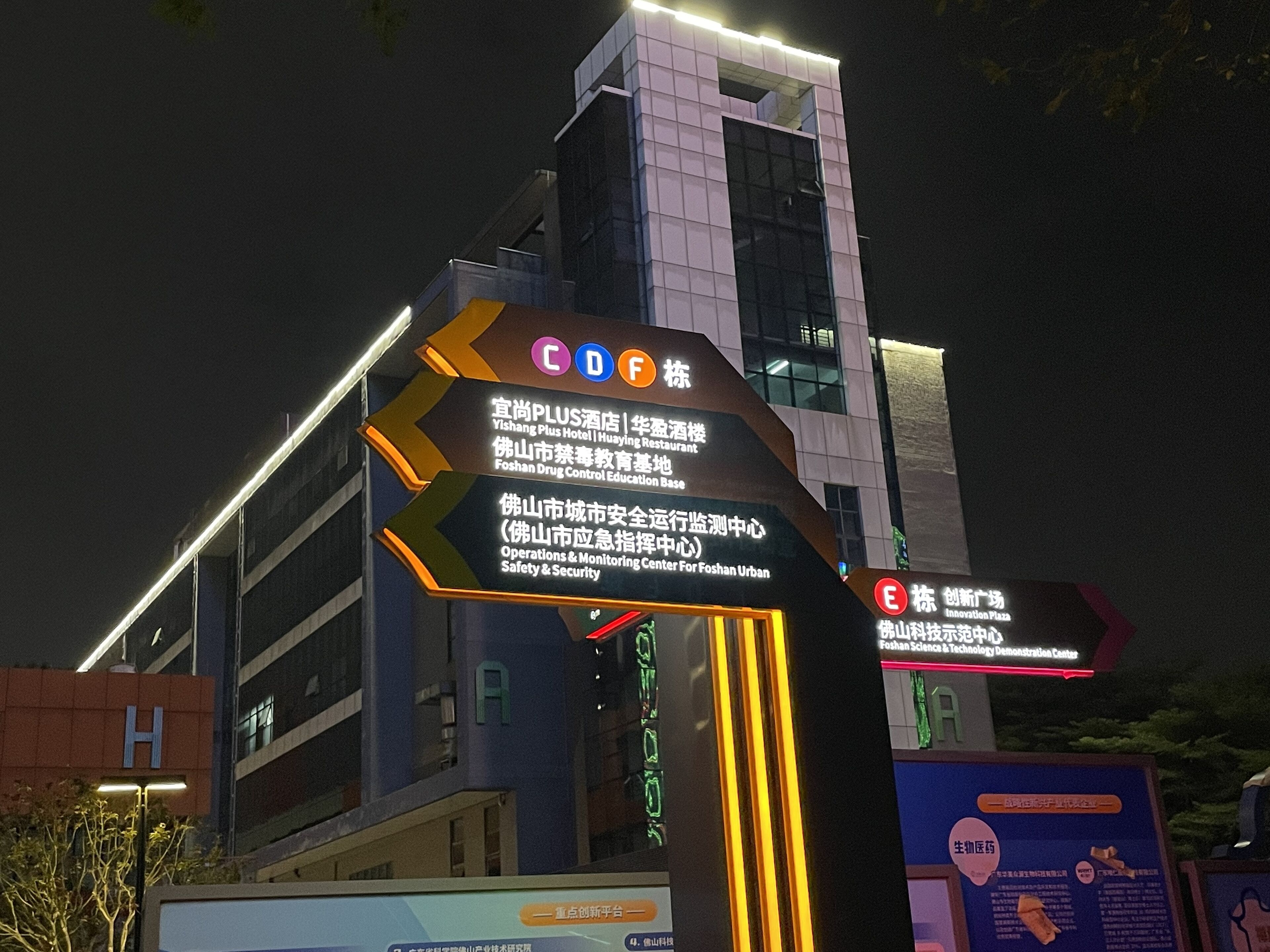 Ein Schild mit verschiedenen Wegweisern und Namen, darunter 'Vishang Plus Hotel' und 'Foshan Drug Control Education Base'.