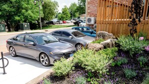 A parking area with two cars, a brick building, and a garden with various plants. - Perth County Inn (Stratford)