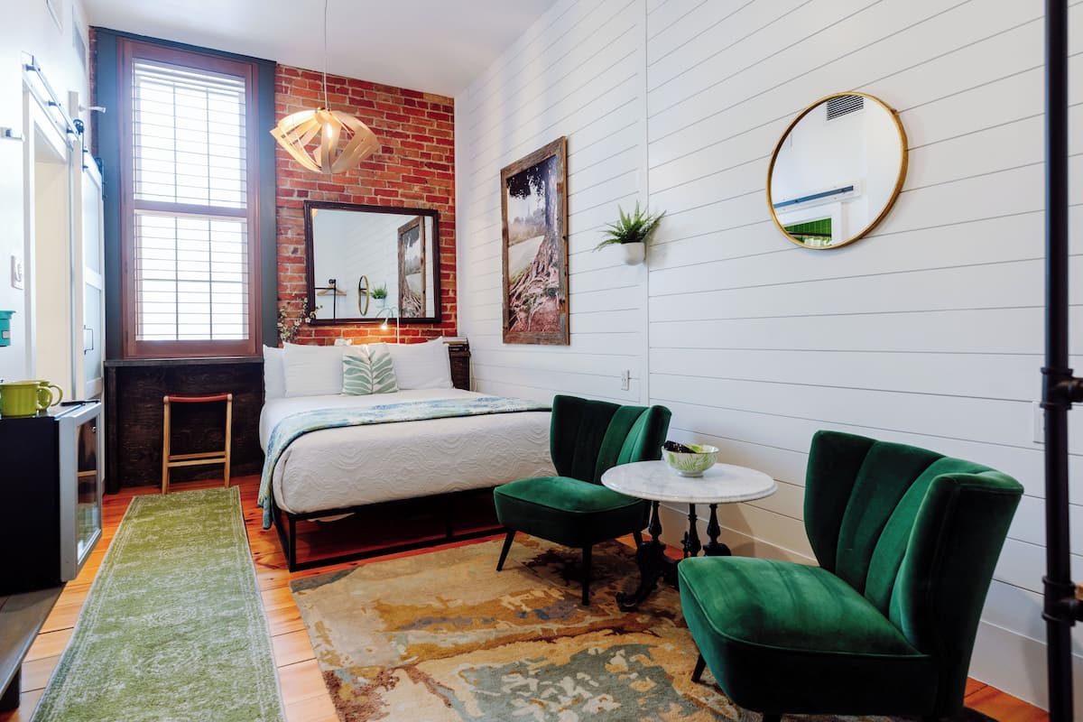 A bedroom with a bed, green armchairs, a small round table, a mirror, and a window with blinds.
