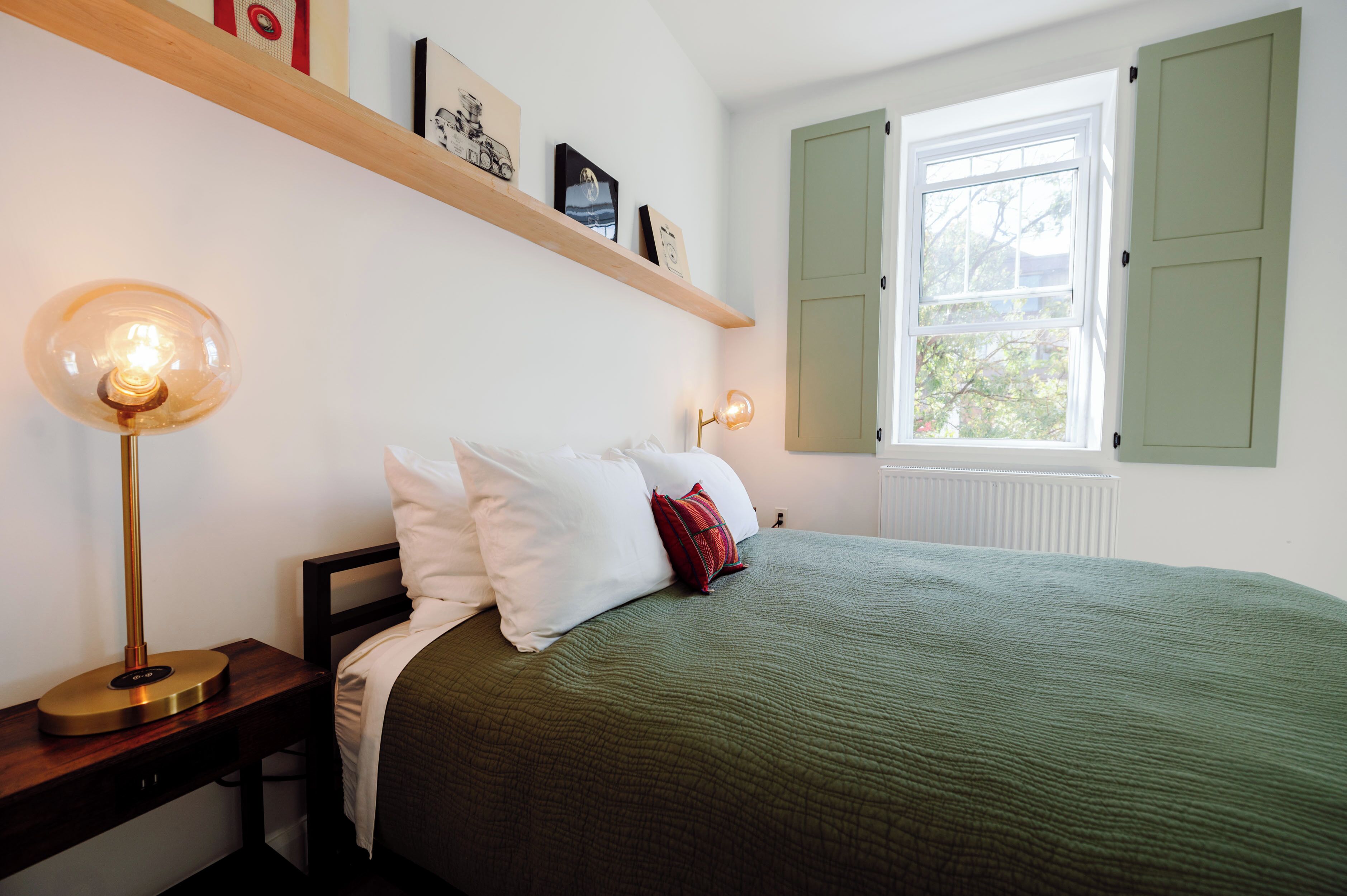 A bedroom with a bed, two bedside lamps, a nightstand, a window with shutters, and a shelf with pictures.