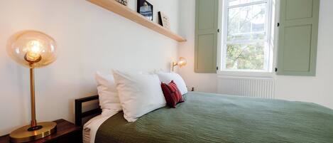 Et soveværelse med en seng, to sengeborde, en lampe på hvert bord, et vindue med skodder og en reol med billeder.