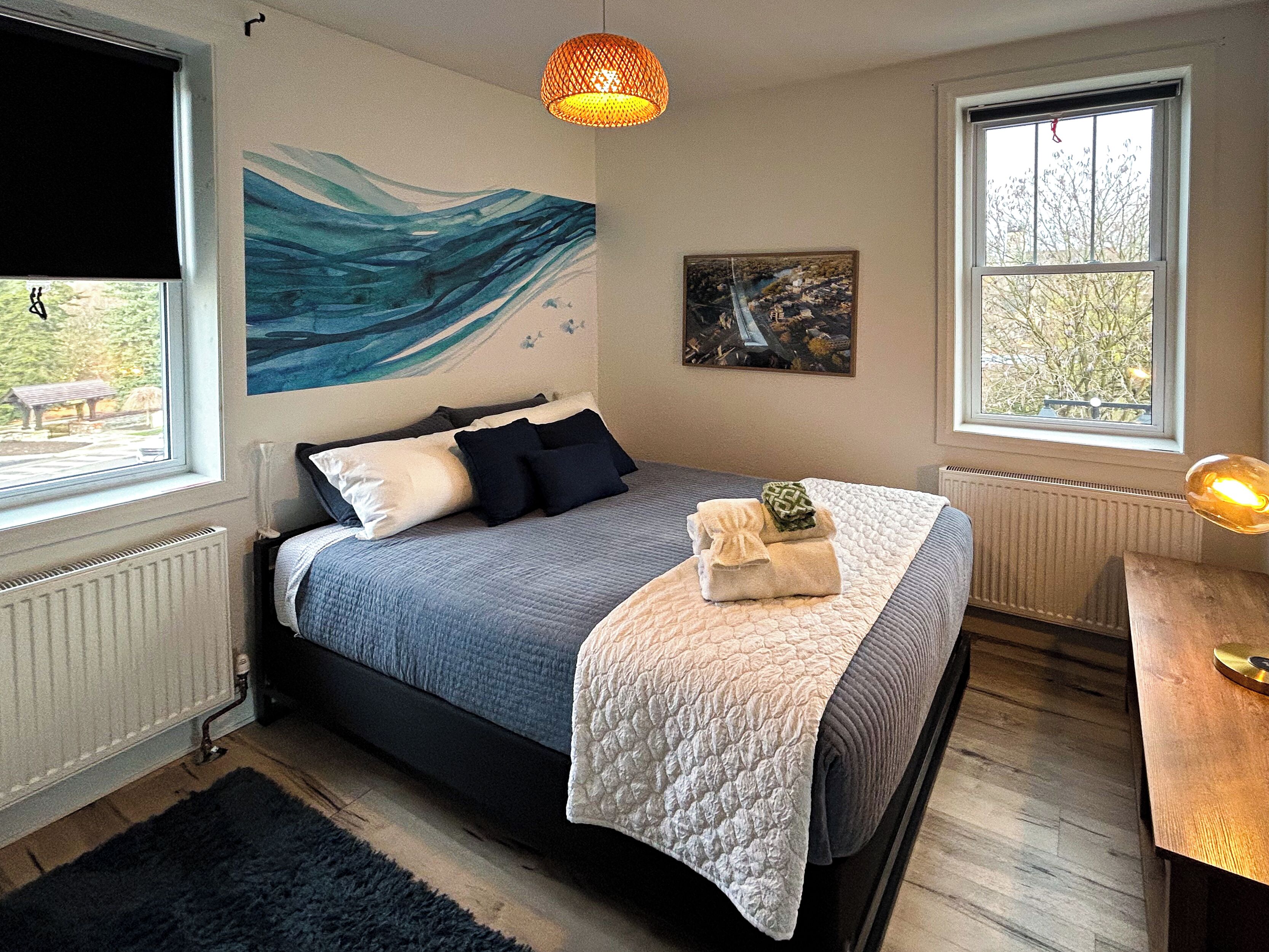 A bedroom with a bed, a large window, a painting, and a lamp.