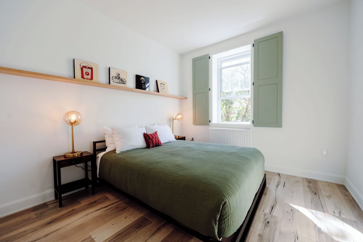 A bedroom with a bed, bedside lamps, a window with shutters, and a shelf with framed pictures.