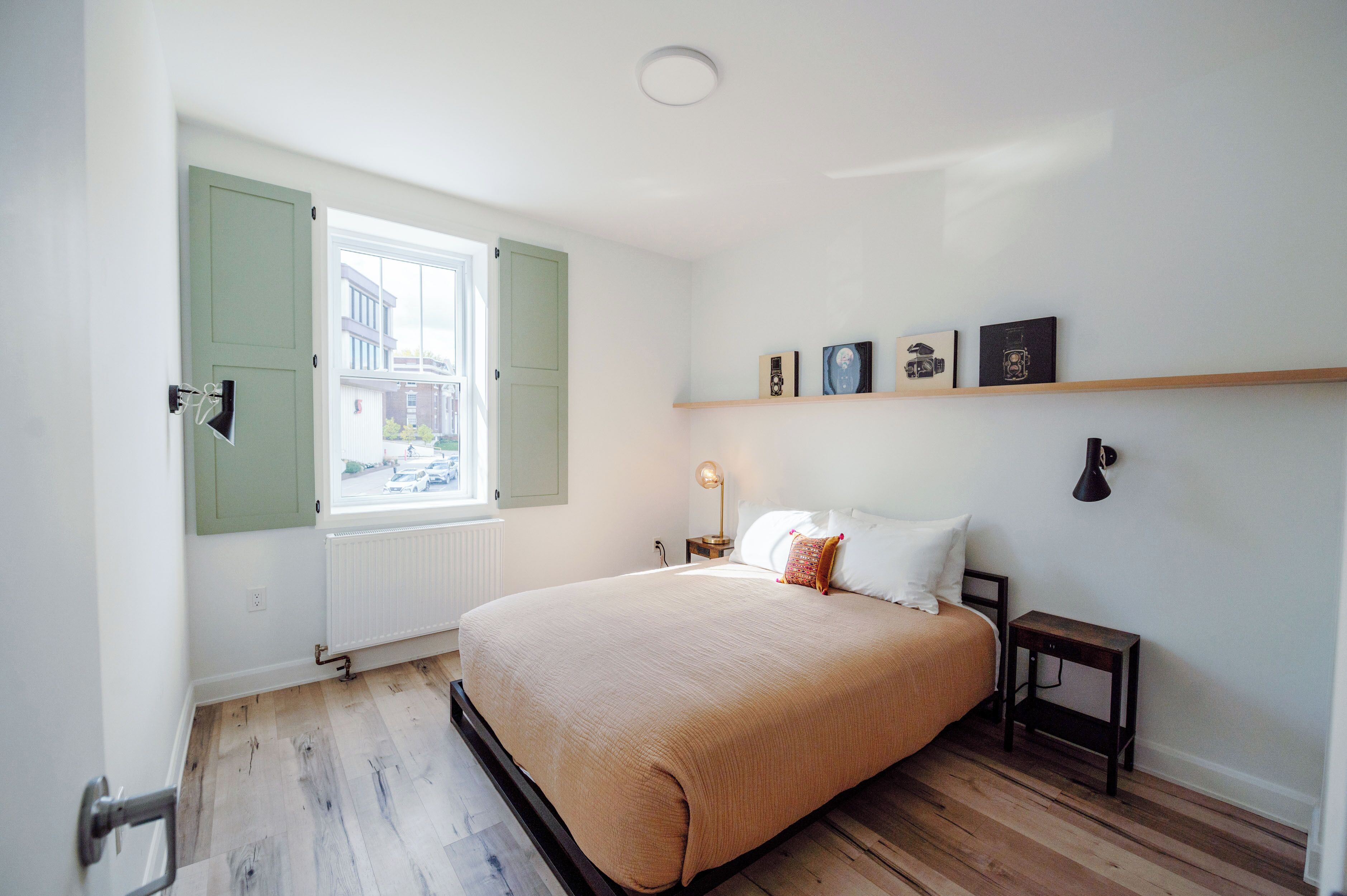 Un dormitorio con cama, ventana con contraventanas, mesita de noche y estante con fotografías.