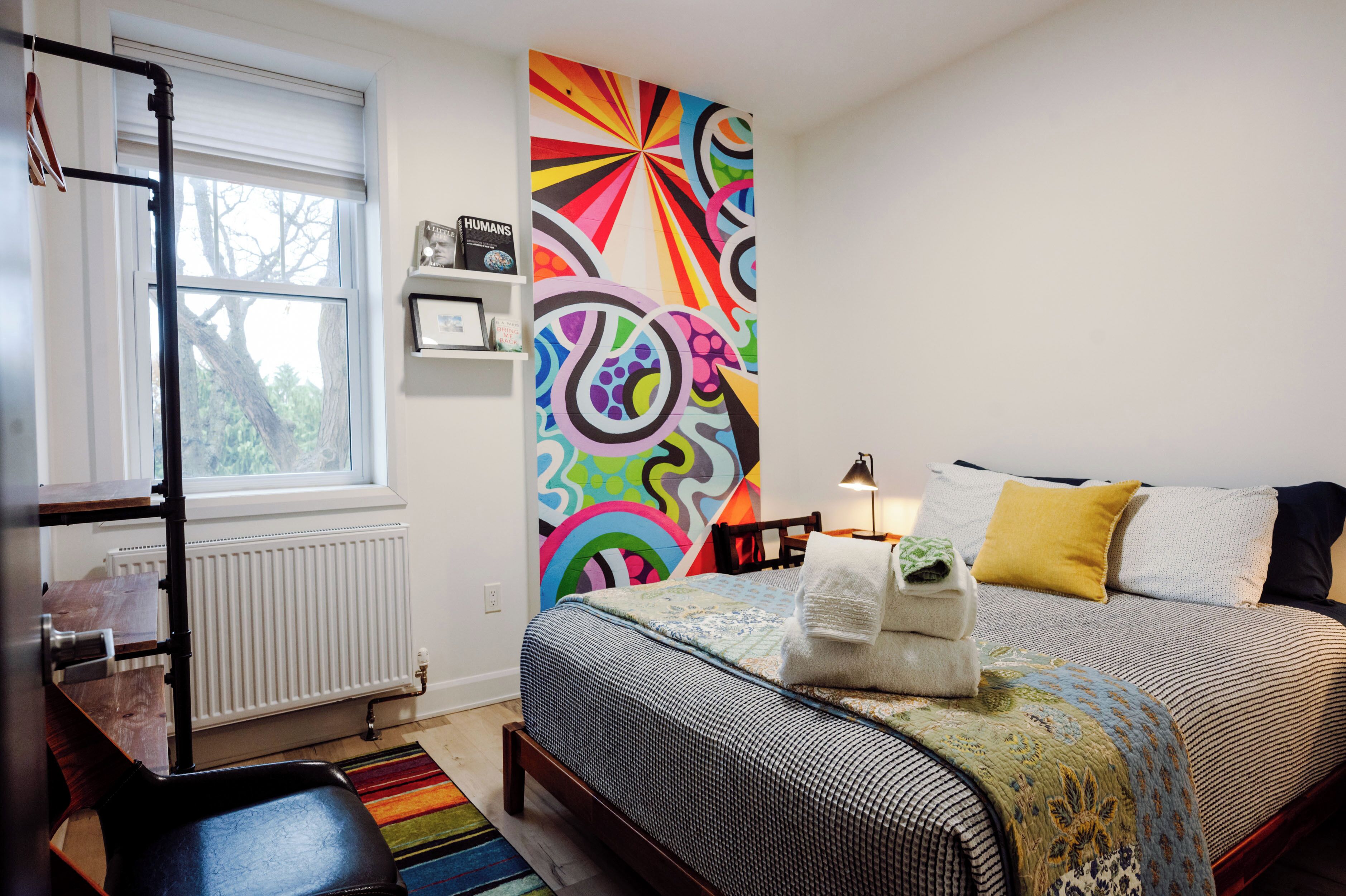 Ein Schlafzimmer mit Bett, Fenster, Heizkörper, einem Regal mit Büchern und einer farbenfrohen, abstrakten Wand.