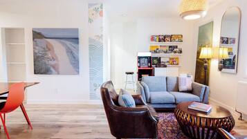 A living room with a sofa, armchair, coffee table, and a dining table with chairs. There is a bookshelf with books and decorative items, a painting on the wall, and a lamp on a side table.