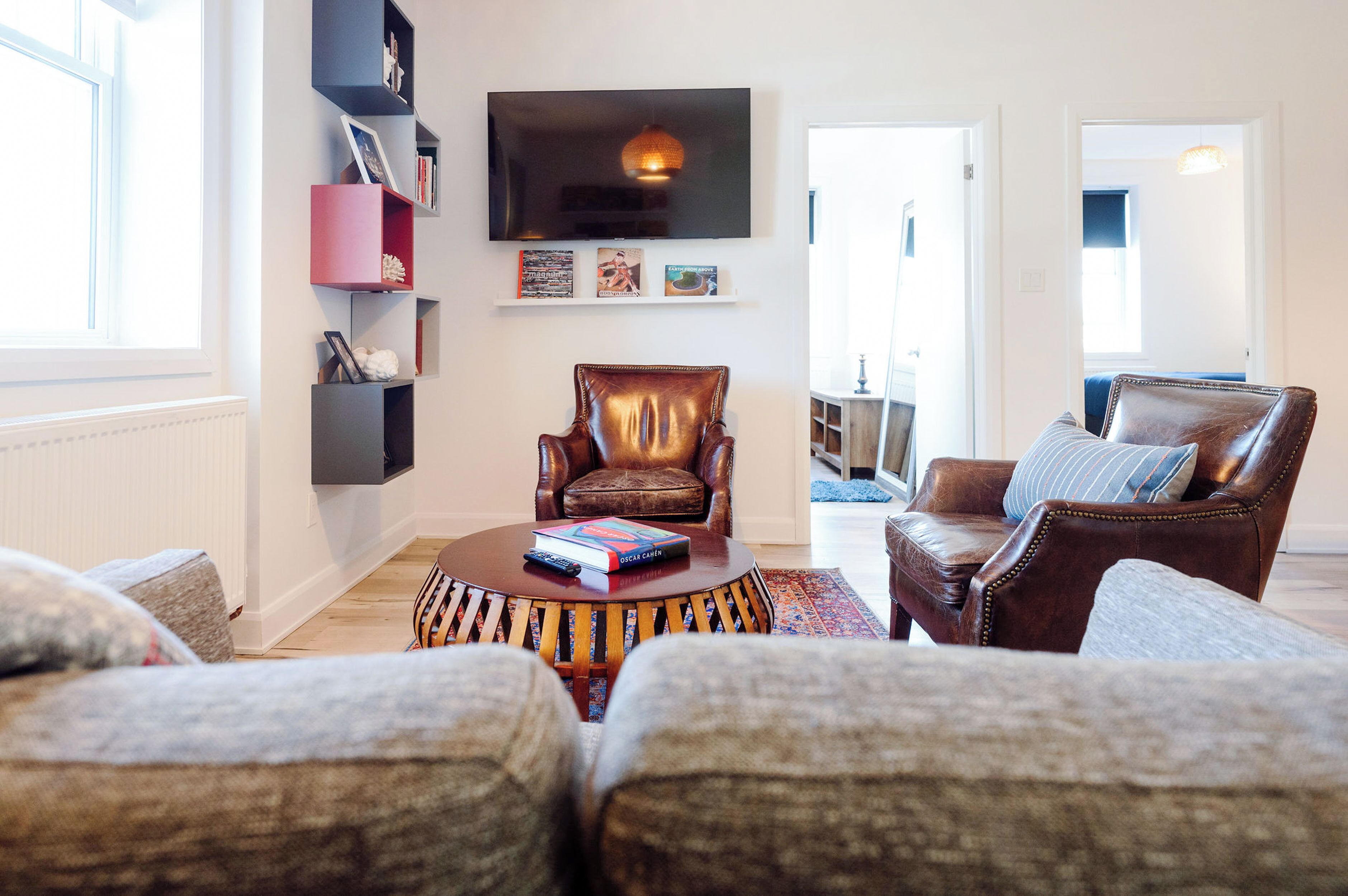 A living room with a brown leather armchair, a round coffee table with books, a flat-screen TV mounted on the wall, and a bookshelf with various items.