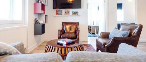 A living room with a brown leather armchair, a round coffee table with books, a flat-screen TV mounted on the wall, and a bookshelf with various items.