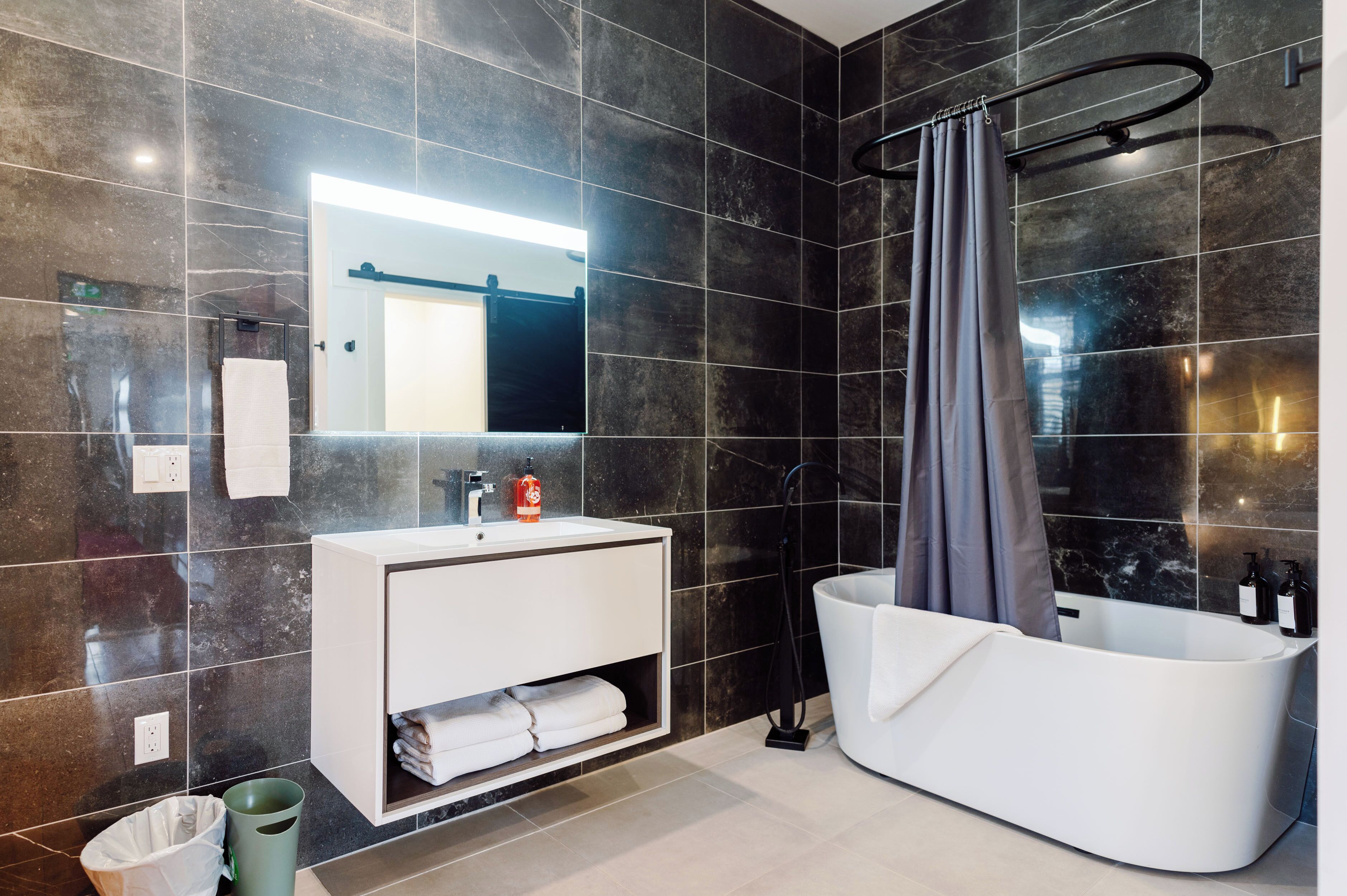 Ein modernes Badezimmer mit dunklen Fliesen, einem weißen Waschtisch, einer großen Badewanne und einer Duschvorhang.