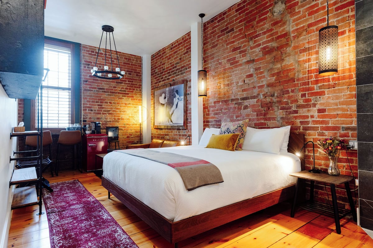 A bedroom with a brick wall, a bed with white bedding, a desk with a chair, and a hanging light fixture.