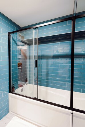 A glass-enclosed shower with a white bathtub, blue tiled walls, and a visible bottle of shampoo.