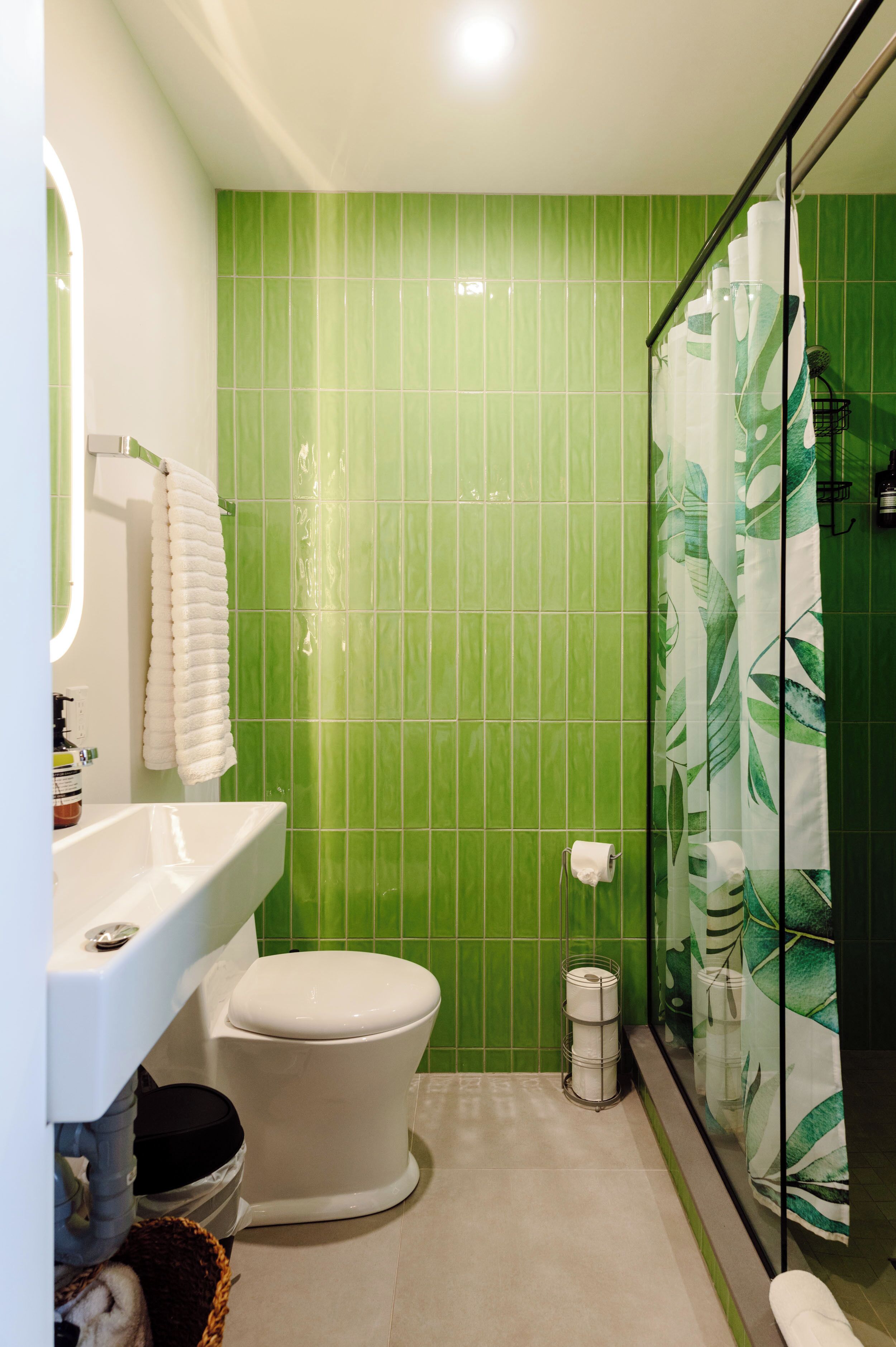 A bathroom with a green tiled wall, a white sink, a toilet, and a shower with a leaf-patterned curtain.
