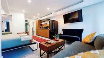 A modern living room with a bed, sofa, coffee table, and a kitchen area with a refrigerator and microwave.