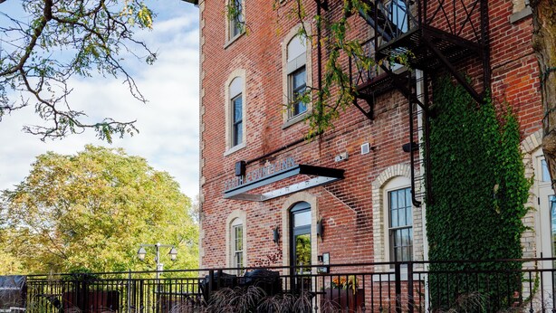 A brick building with a fire escape, surrounded by greenery and flowering plants. - Perth County Inn (Stratford)