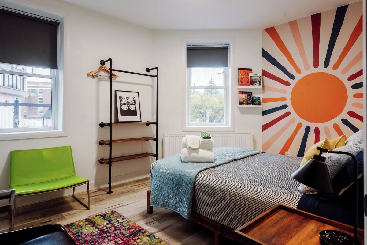 A bedroom with a bed, a green chair, a wooden shelf, a radiator, and a large sun mural on the wall.