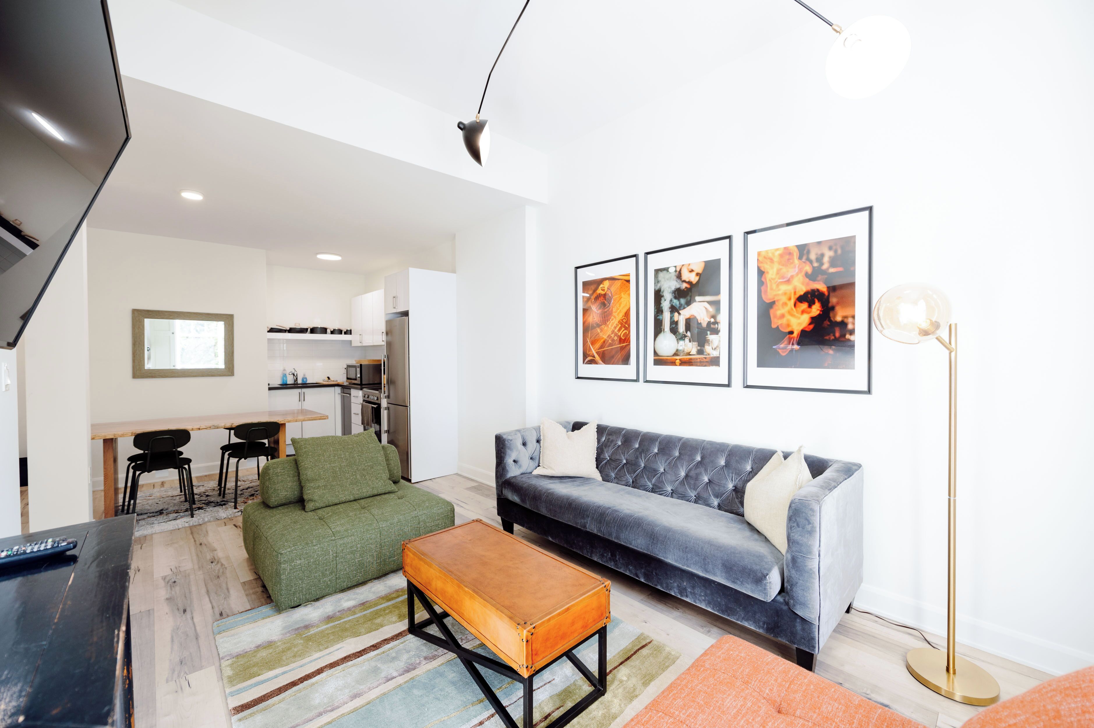 A modern living room with a grey sofa, a green armchair, and a wooden coffee table. There is a kitchen in the background with a dining table and chairs.