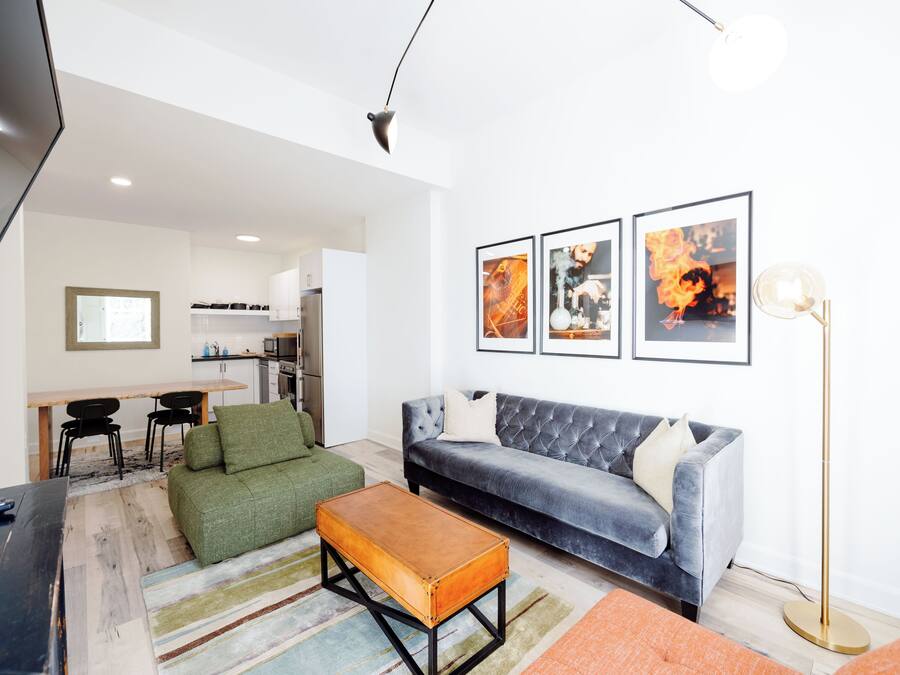 A modern living room with a grey sofa, a green armchair, and a wooden coffee table. There is a kitchen in the background with a dining table and chairs.