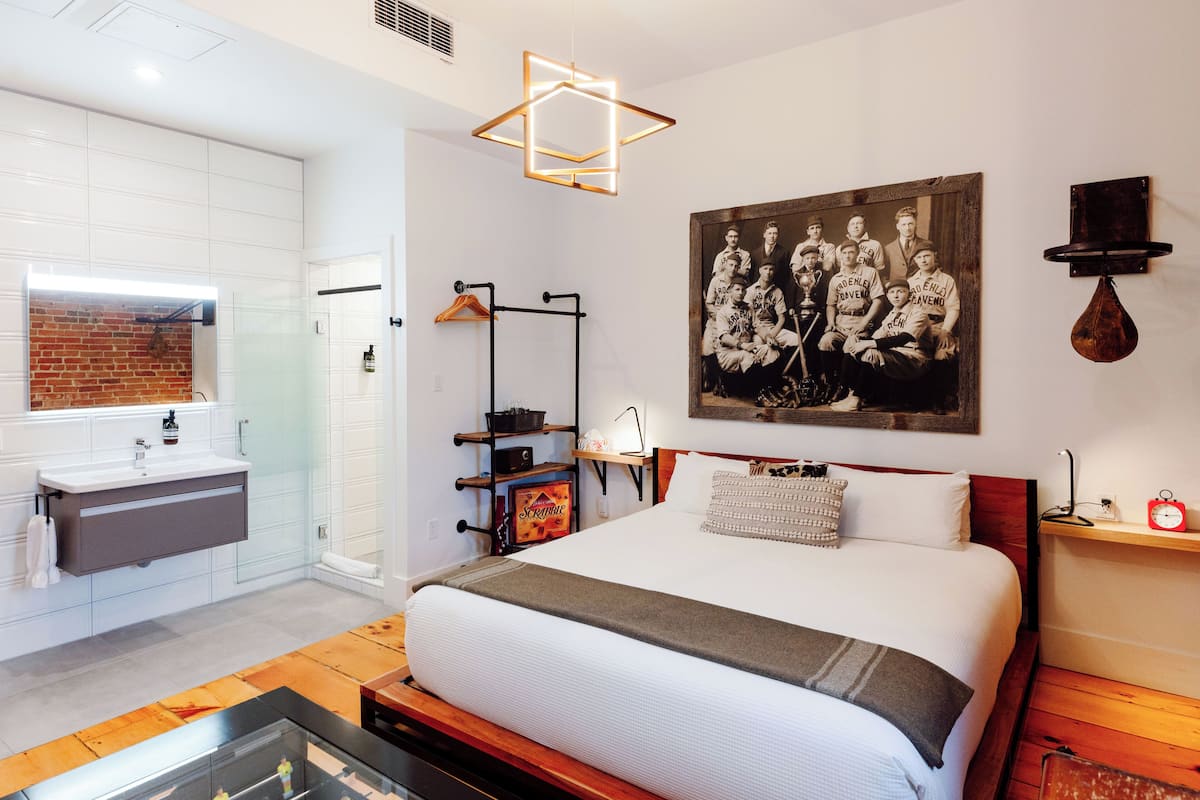 A modern hotel room with a large bed, a bathroom with a glass shower enclosure, a wooden floor, and a wall-mounted picture.