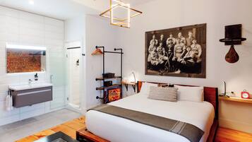Una camera d'albergo moderna con un letto grande, bagno con box doccia in vetro, pavimento in legno e un quadro appeso al muro.