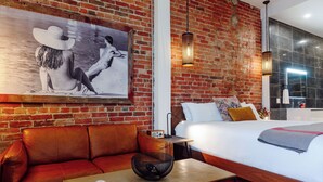 A hotel room with a brick wall, a large bed, a leather sofa, and a painting of a woman in a hat. - Perth County Inn (Stratford)