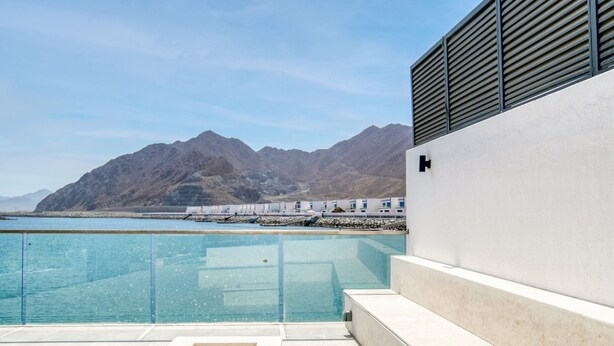 Ein moderner Poolbereich mit klarem, blauem Wasser, einer Steinsitzbank und Blick auf Berge sowie ein Gebäude an der Küste im Hintergrund.