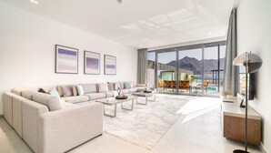 Ein modernes Wohnzimmer mit einer großen Couch, einem Couchtisch und einem Essbereich mit Stühlen. An den Wänden hängen gerahmte Bilder, eine Stehlampe und durch große Fenster ist eine Berglandschaft zu sehen.