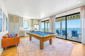 A pool table in a room with a view of the sea, a sofa, and a kitchen area.