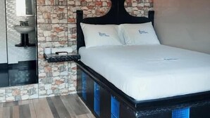 Une chambre avec un mur décoré d’un motif en pierre, un lit avec une literie blanche et une table de chevet dotée d’un miroir et d’un petit lavabo.