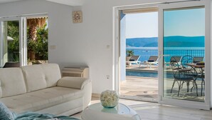 A modern living room with a beige sofa, a glass coffee table with a vase, and a view of a pool and lounge chairs on a balcony. - Seafront Villa Azzurro with heated pool, panoramic sea views, walking distance to beach-shop-restaurant (Stanici)