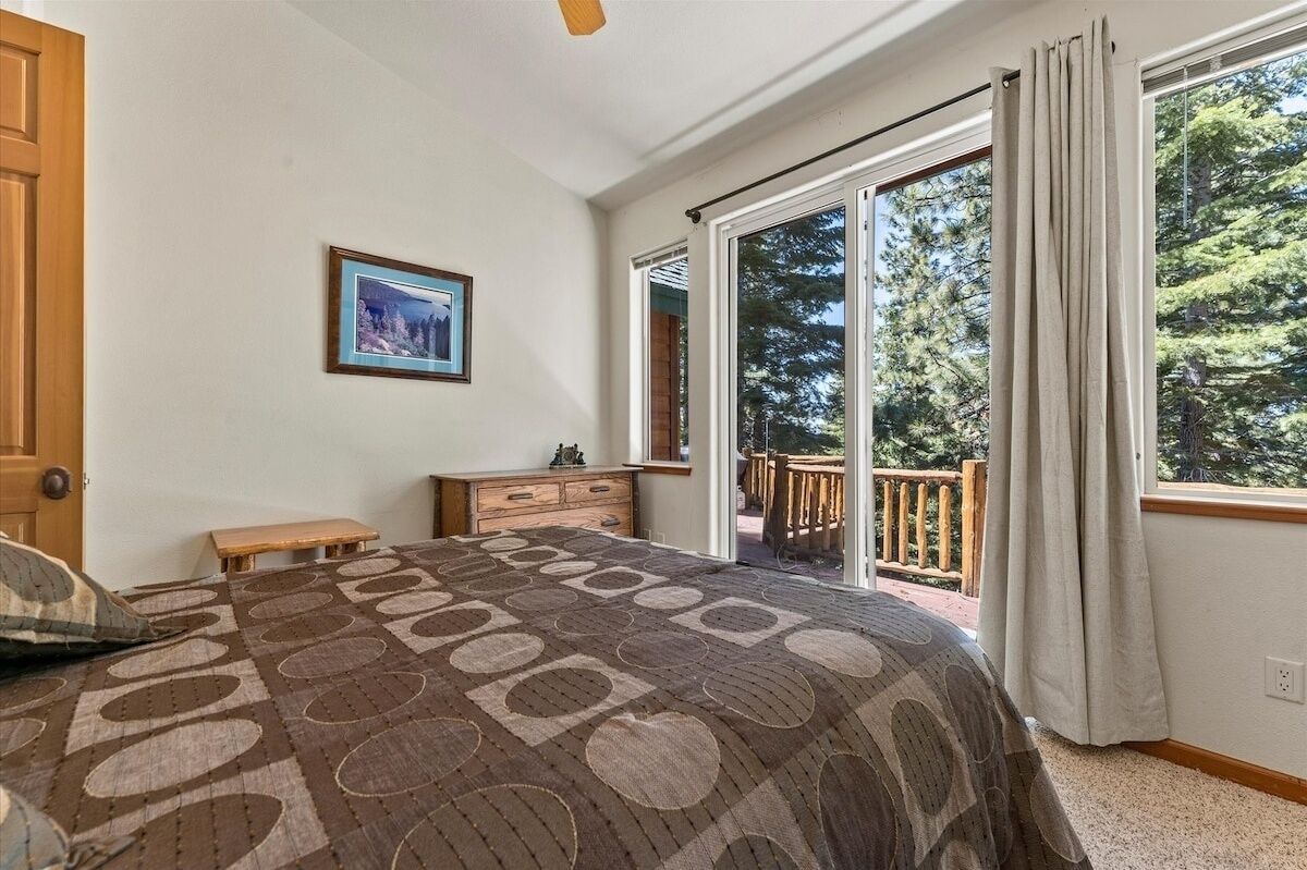 A bedroom with a large bed, a dresser, a bench, a framed picture, and a sliding glass door leading to a balcony with a view of trees.