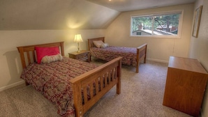 Quarto com duas camas de madeira, uma mesa de cabeceira, abajur, janela e cómoda de madeira.
