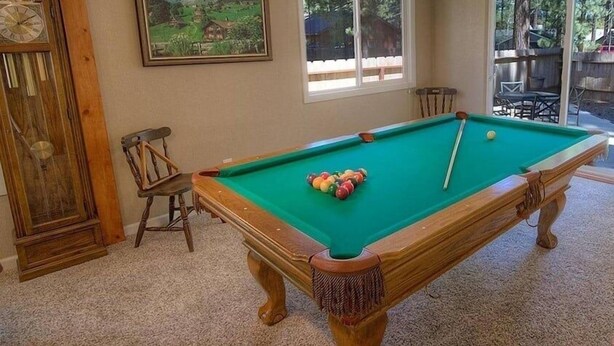 Uma mesa de bilhar com bolas, um relógio e uma pintura em um quarto com pisos carpetados e vigas de madeira.