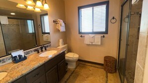 A bathroom with a double sink, a toilet, a shower, and a window with blinds.
