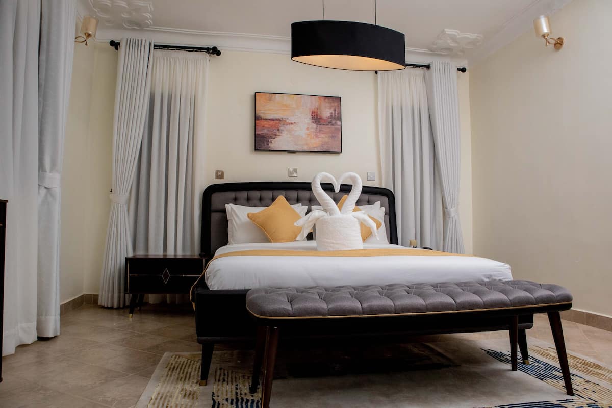 Un dormitorio con cama, mesita de noche, banco, un cuadro y cortinas.