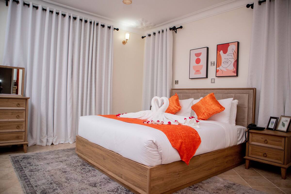 Una cama bien hecha con ropa de cama blanca y detalles naranja, una mesita de noche de madera con un cuadro enmarcado, y un ventanal con cortinas blancas.