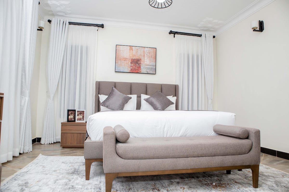 Un dormitorio con cama, una banca, una mesita de noche y un cuadro en la pared.