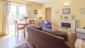 A living room with a sofa, a dining table with chairs, a fireplace, and a view through a window with curtains. - Curlew Cottage (Hexham)
