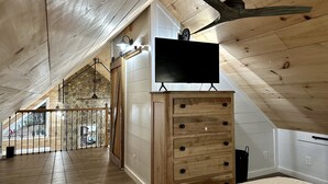 A spacious attic room with a wooden ceiling, a large TV on a wooden dresser, and a view of the outdoors through a window.