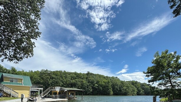 A wooden dock with two Adirondack chairs facing a lake, surrounded by trees and a building with a green roof.