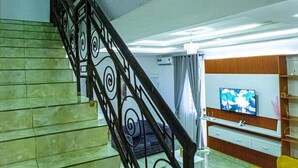 Un escalier avec une rampe noire et des marches en marbre vert menant à un salon équipé d’un téléviseur, d’un canapé jaune et de panneaux de bois.