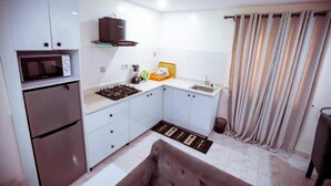 A compact kitchen with a microwave, stove, and sink. There is a small countertop with a coffee maker and a trash bin. The room has a window with curtains.