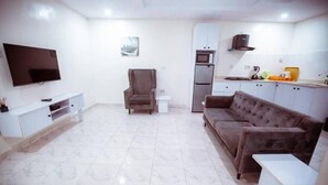 A living room with a flat-screen TV, a brown sofa, a white coffee table, a white cabinet, and a kitchen area with a microwave and refrigerator.