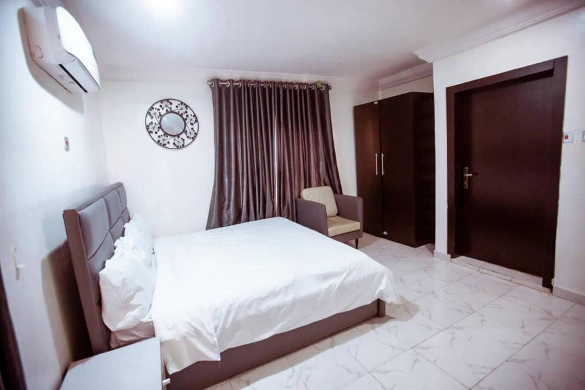 Quarto de hotel com cama, uma cadeira, um guarda-roupa e um espelho na parede.
