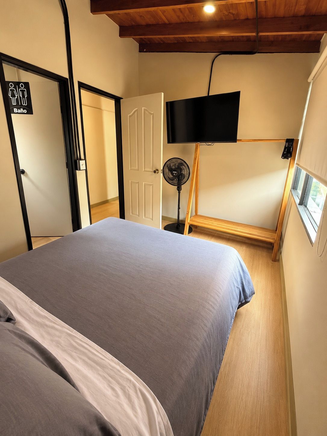 A bedroom with a bed, a TV mounted on the wall, a fan, and a door with a sign indicating a bathroom.