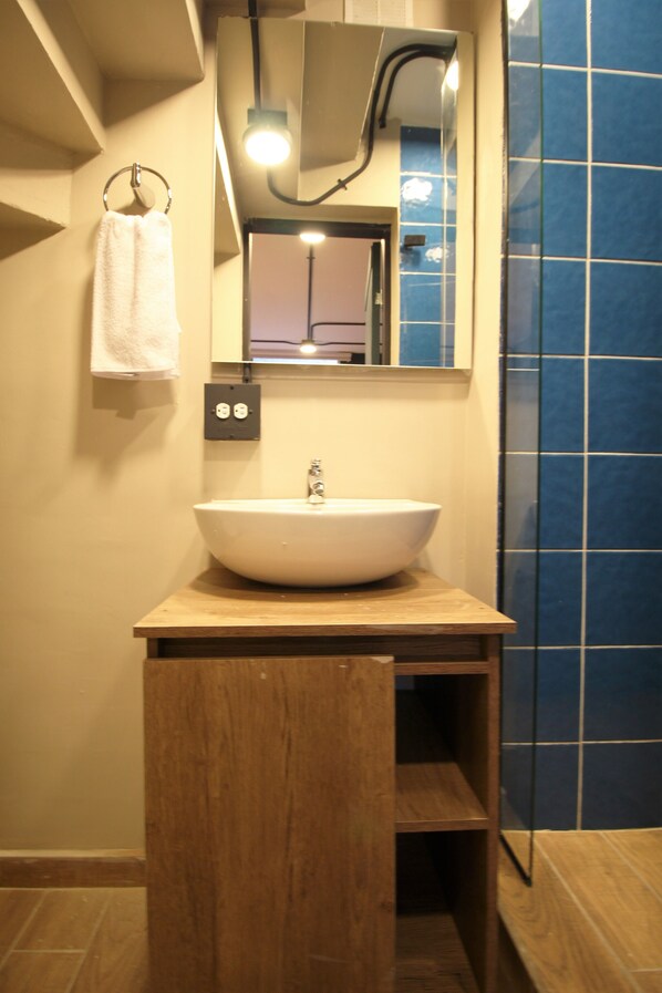 A bathroom with a wooden vanity, a white sink, a towel hanging on a metal ring, a mirror, and blue-tiled walls. - One Living by Roots Rentals (Medellín)
