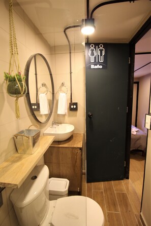 A bathroom with a toilet, sink, and mirror. There is a door with a sign indicating a men's and women's bathroom. - One Living by Roots Rentals (Medellín)