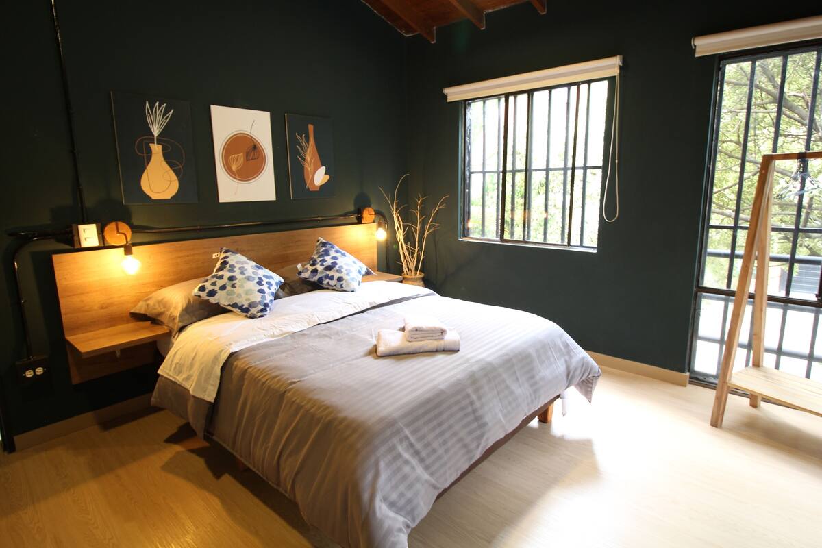 Une chambre à coucher avec un lit, des tables de chevet, une fenêtre avec des barreaux et des œuvres d’art accrochées au mur.