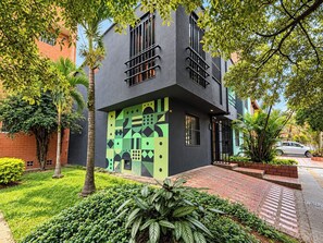 A modern two-story building with a geometric facade, surrounded by lush greenery and a paved walkway. - One Living by Roots Rentals (Medellín)